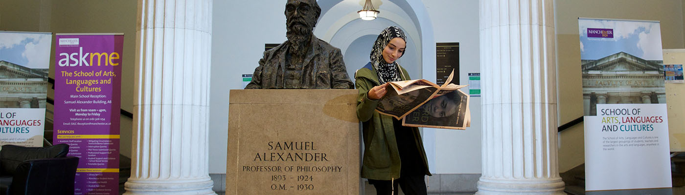 Female student in headscarf reading newspaper leaning on the Samuel Alexander bust in the foyer